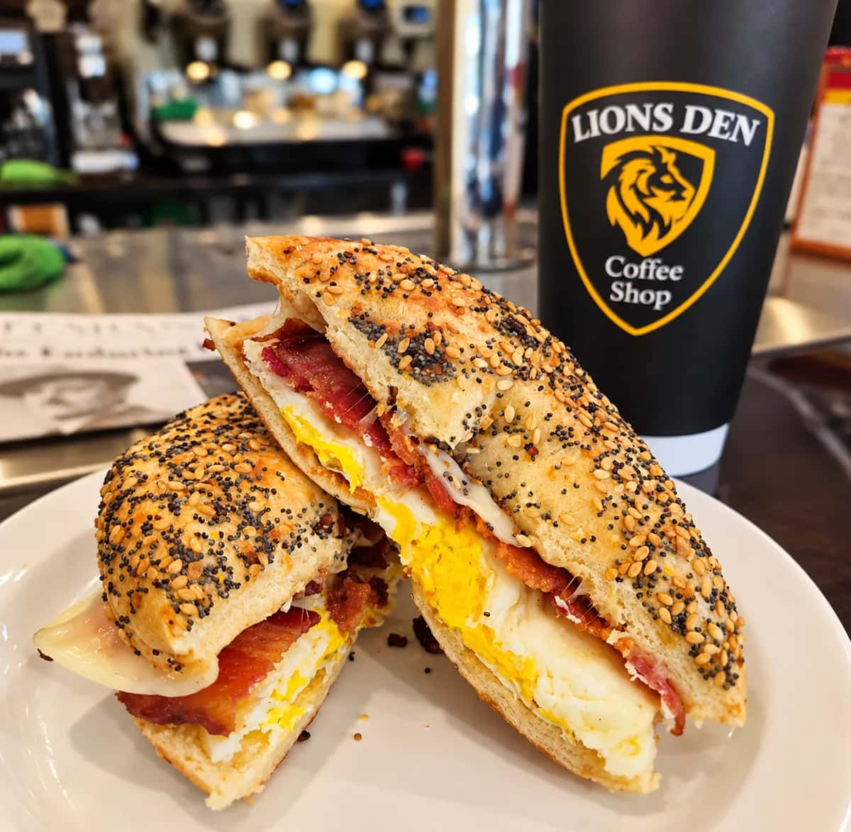 Bacon, egg and cheese on an everything bagel, cut in half on a white plate, with a Lions Den Coffee Shop cup behind it
