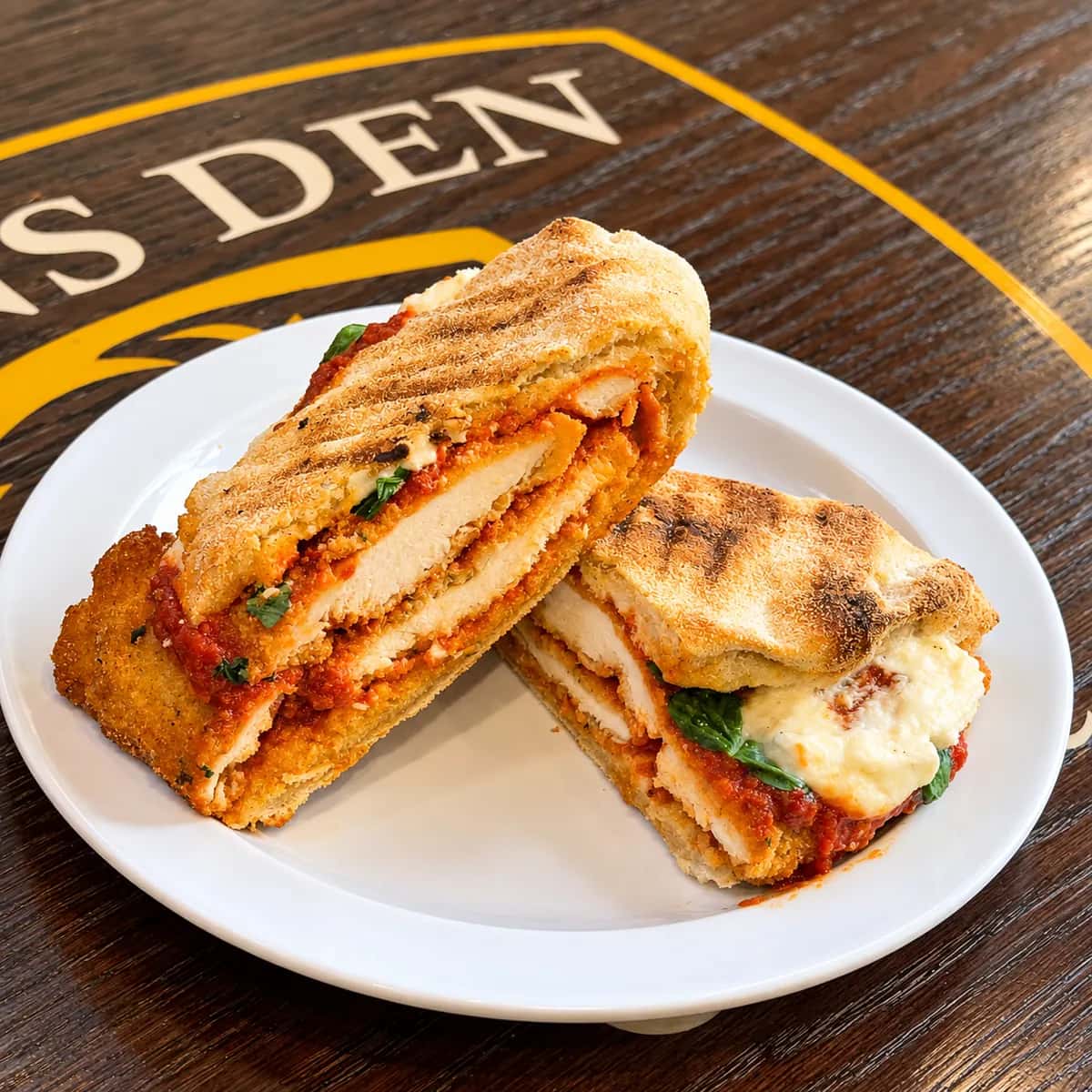 Chicken parm panini cut in half, showing breaded chicken, red sauce, ricotta, and basil on a Lions Den Coffee Shop table.