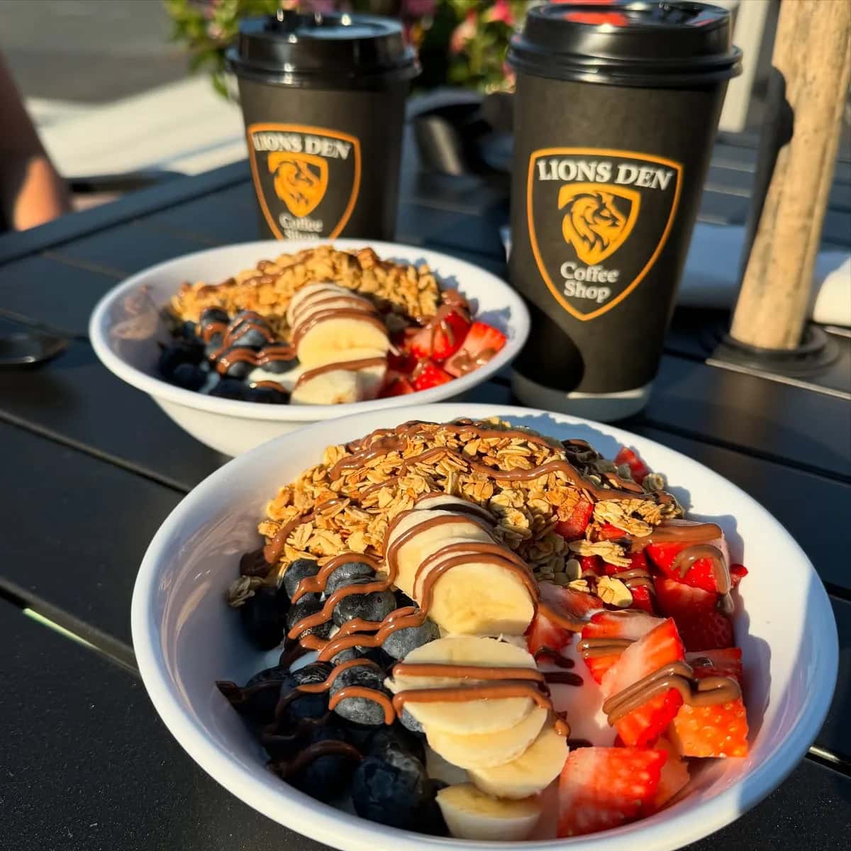 Two parfait bowls topped with granola, blueberries, sliced banana, strawberries and a drizzle, beside Lions Den Coffee Shop cups on a sunny patio table