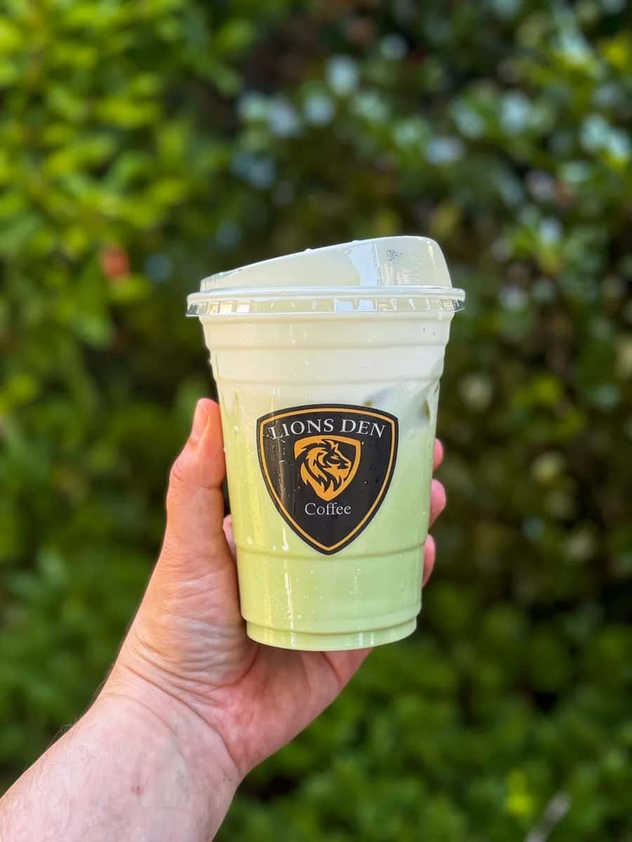 Hand holding an iced green matcha drink in a Lions Den Coffee cup against green hedges