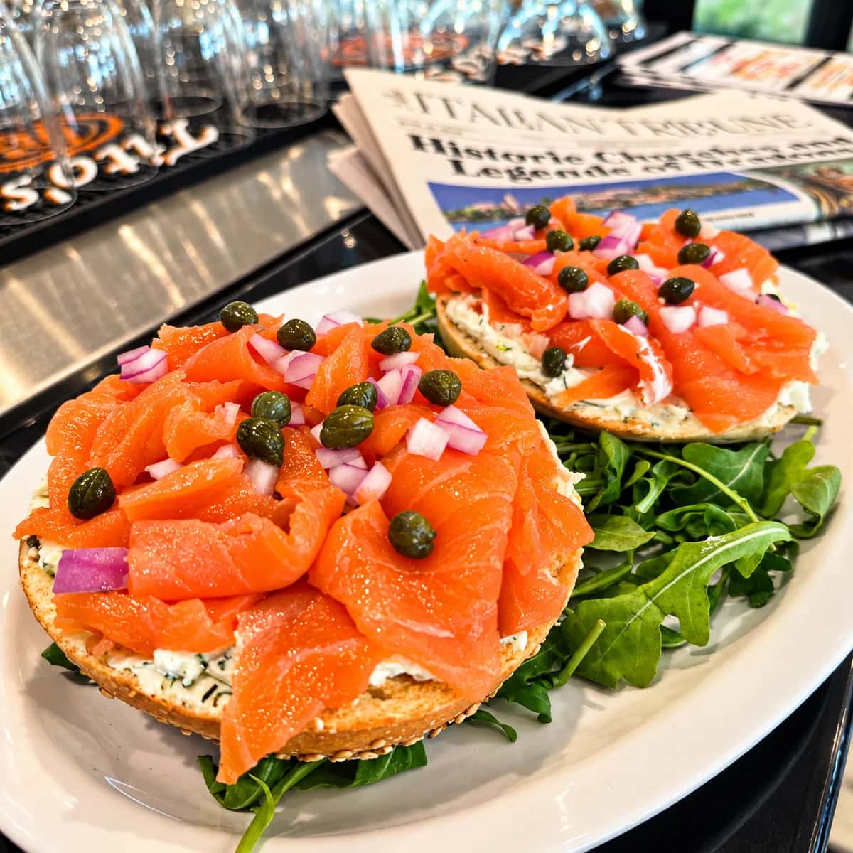 Two open bagel halves spread with herbed cream cheese and topped with smoked salmon, capers and diced red onion, on a plate with arugula