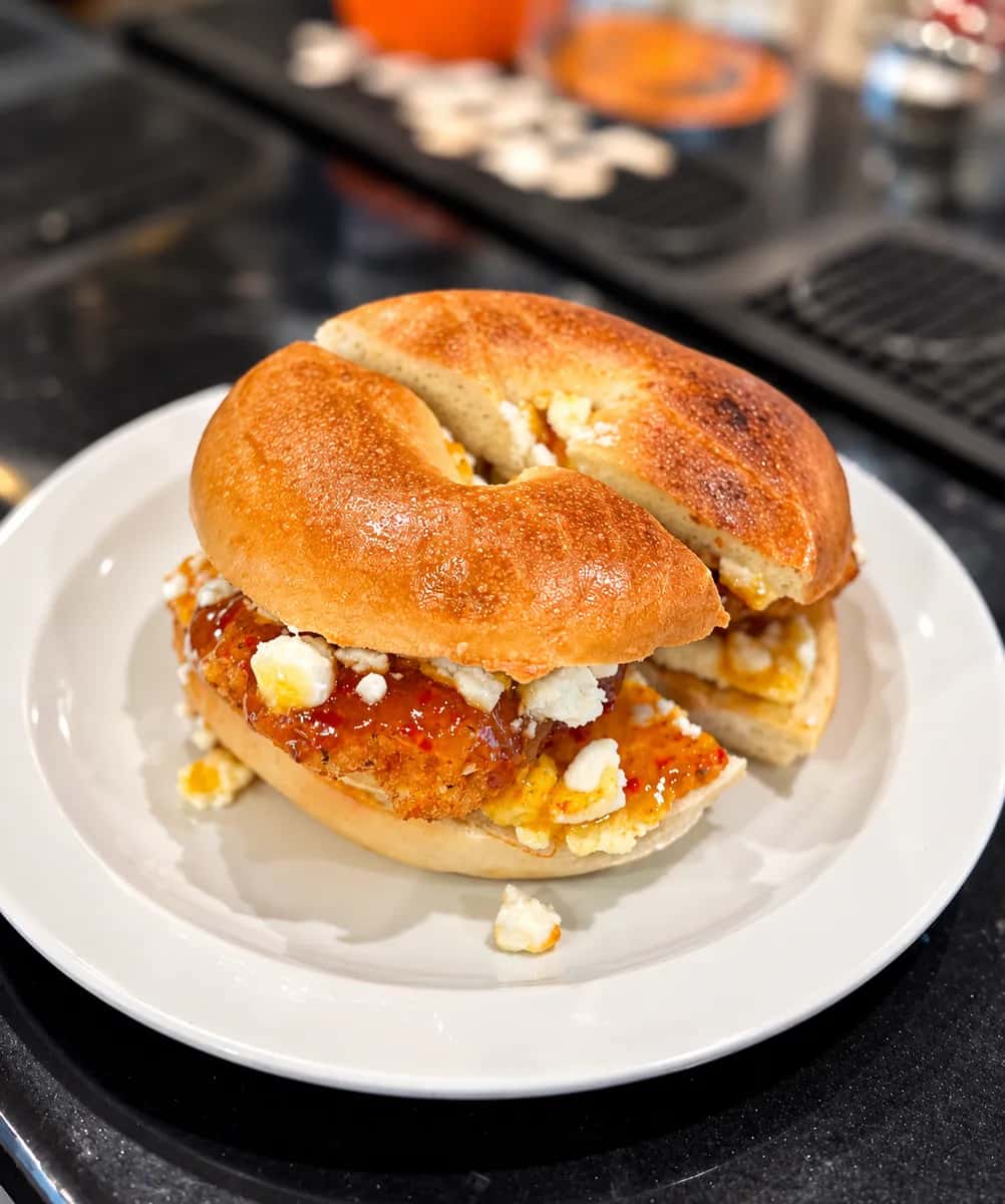 Bagel sandwich with a breaded chicken cutlet, red pepper jelly and crumbled white cheese on a white plate