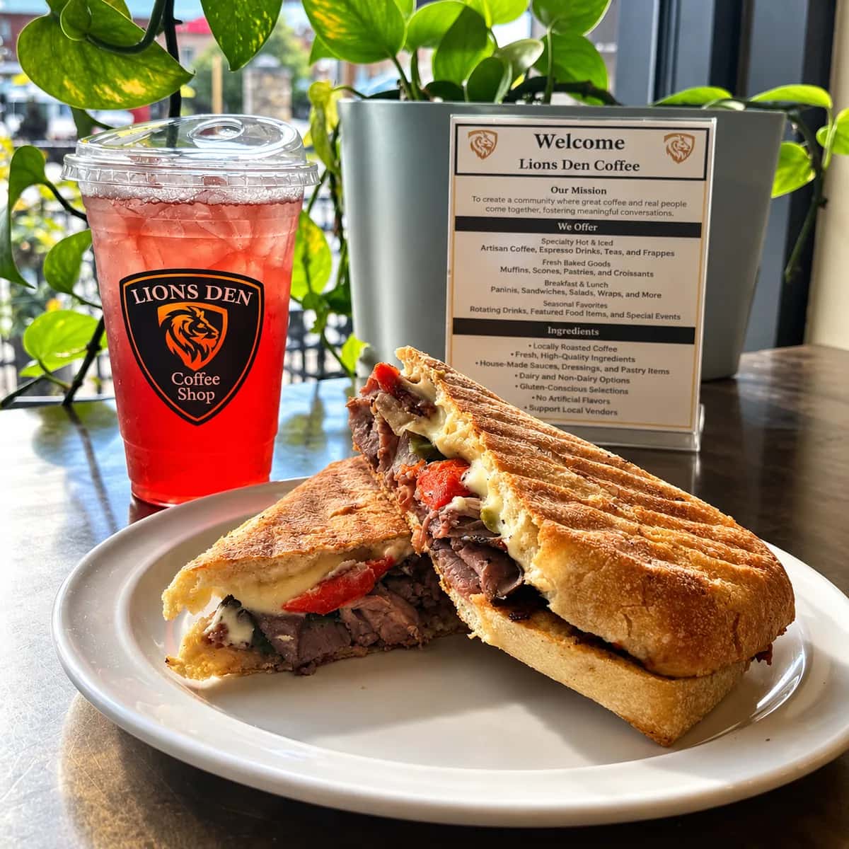 Grilled panini cut in half, showing sliced roast beef, red peppers and melted cheese, beside a red iced drink in a Lions Den Coffee Shop cup