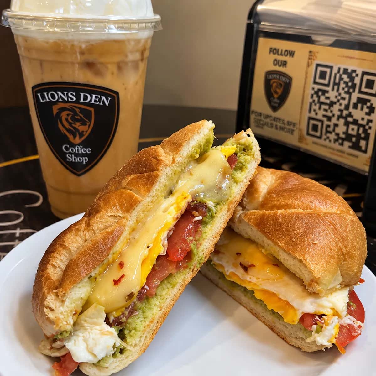 Breakfast sandwich cut in half, showing avocado spread, tomato, egg and melted cheese, with an iced coffee behind it